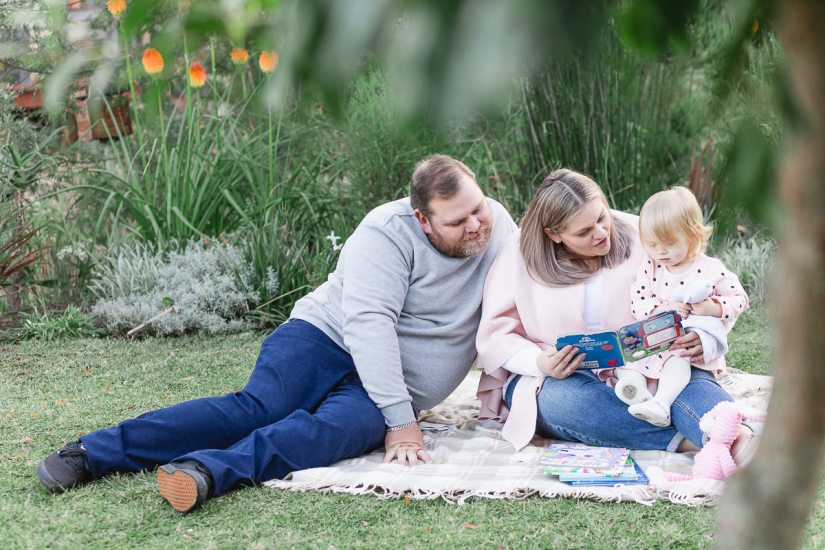 Mom, Dad and baby girl sitting on blanket while reading a childs book blurred out greenery and flowers at Woodlands self catering accommodation in the background during a lifestyle family photo shoot by a Sedgefield photographer moi du toi photography
