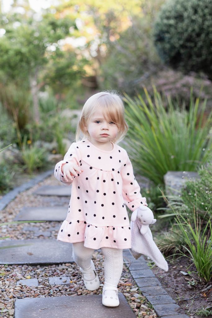 Full length shot of Toddler dressed in pink walking down path towards the camera, blurred out greenery at Woodlands self catering accommodation in the background during a lifestyle family photo shoot by a Sedgefield photographer moi du toi photography