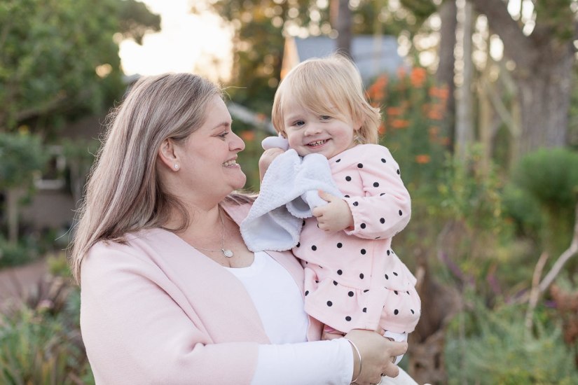 Mom holding and smiling at baby daughter while she laughs, blurred out greenery and setting sun at Woodlands self catering accommodation in the background during a lifestyle family photo shoot by a Sedgefield photographer moi du toi photography