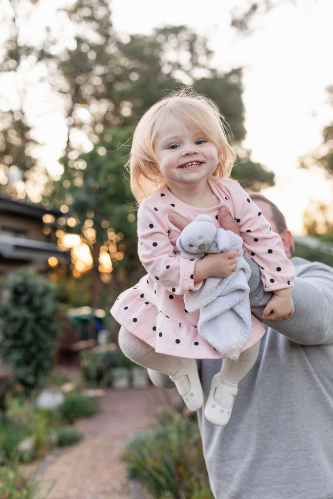 Dad playing airplane game while holding his baby daughter while she laughs, burred out greenery and sunset at Woodlands self catering accommodation in the background during a lifestyle family photo shoot by a Sedgefield photographer moi du toi photography