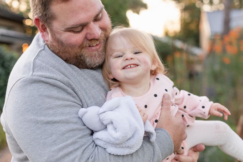 Dad holding his baby daughter while she laughs, both looking directly at the camera blurred out greenery and flowers at Woodlands self catering accommodation in the background during a lifestyle family photo shoot by a Sedgefield photographer moi du toi photography