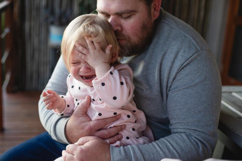 Dad cuddling his baby daughter while she cries during a lifestyle family photo shoot by a Sedgefield photographer moi du toi photography