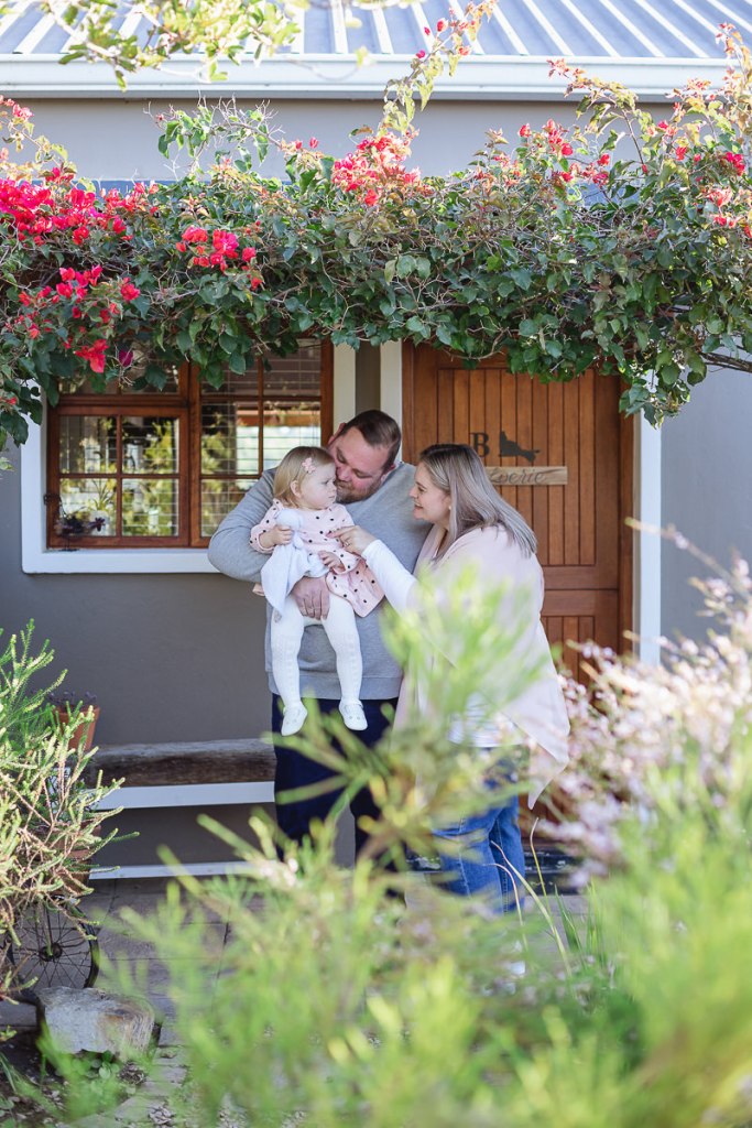 Dad holding his baby daughter while Mom interacts with her outside their cottage door at Woodlands self catering accommodation in Knysna during a lifestyle family photo shoot by a Sedgefield photographer moi du toi photography