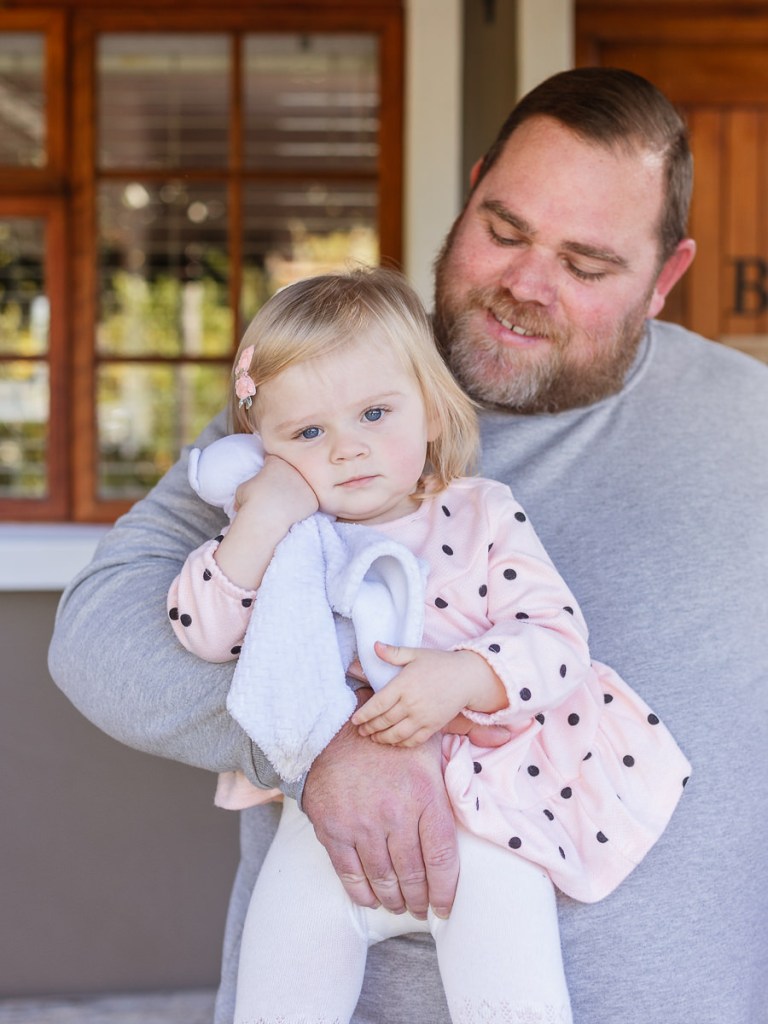 Dad holding his baby daughter cuddling a soft toy outside their cottage door at Woodlands self catering accommodation in Knysna during a lifestyle family photo shoot by a Sedgefield photographer moi du toi photography