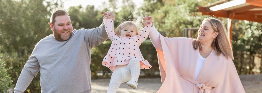 Mom & Dad swinging their female toddler between them while she laughs with blurred out greenery and flowers at Woodlands self catering accommodation in the background during a lifestyle family photo shoot by a Sedgefield photographer moi du toi photography