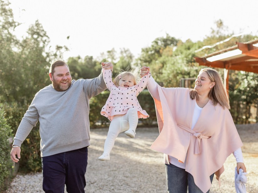 Mom & Dad swinging their female toddler between them while she laughs with blurred out greenery and flowers at Woodlands self catering accommodation in the background during a lifestyle family photo shoot by a Sedgefield photographer moi du toi photography
