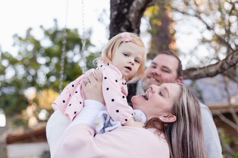 Mom holding and smiling at baby daughter, Dad watches on background blurred out greenery at Woodlands self catering accommodation in the background during a lifestyle family photo shoot by a Sedgefield photographer moi du toi photography